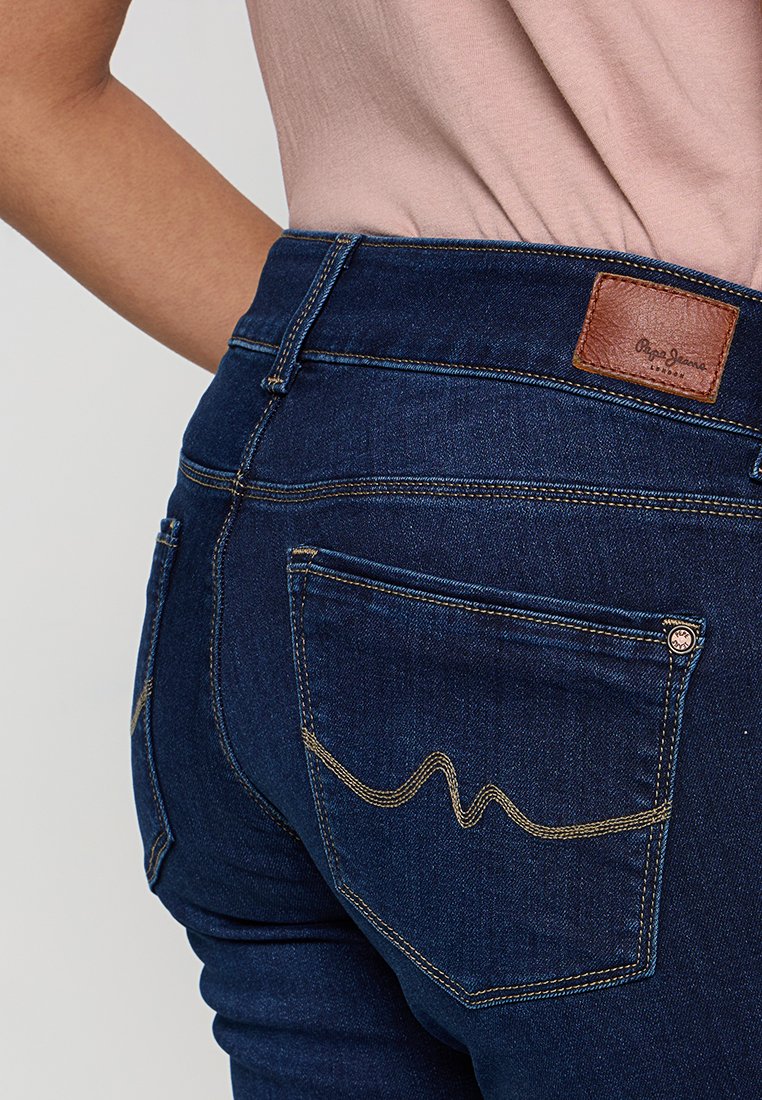 Dunkelblaue Jeans mit hoher Taille, ausgestattet mit einem braunen Lederpatch und goldenen Nähten an den Hintertaschen, die ein wellenförmiges Muster aufweisen.