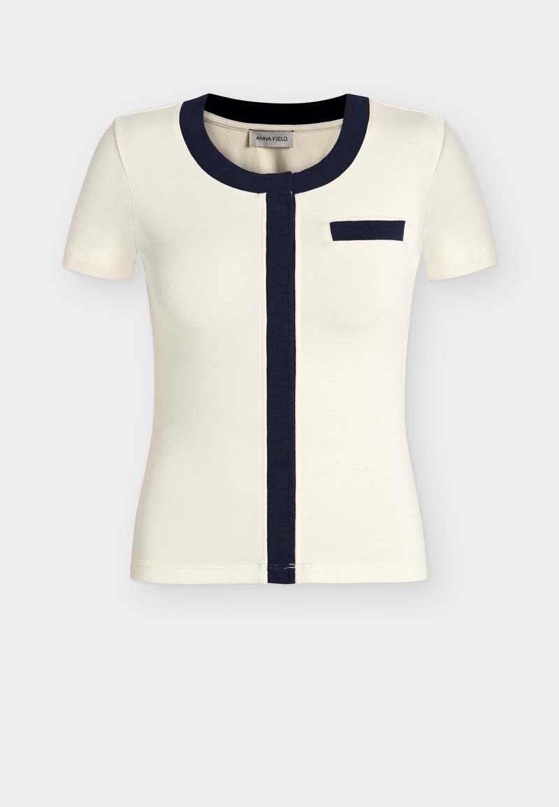 Μπλούζα με κοντά μανίκια σε κρεμ χρώμα με στρογγυλή λαιμόκοψη σε ναυτικό μπλε, κάθετη κεντρική ρίγα και μικρή τσέπη στο στήθος.