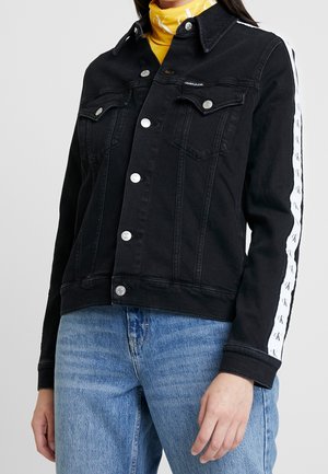 Femme portant une veste en jean noire avec des bandes blanches avec logo sur les manches, des boutons argentés, un col roulé jaune et un jean bleu.
