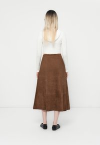 Jupe midi en daim marron avec un design évasé et des détails de coutures invisibles, associée à un haut ajusté à manches longues blanc et des chaussures noires.