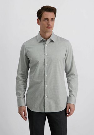 Mann trägt ein hellgraues, langärmliges Hemd mit Knöpfen und Kragen, steht vor einem schlichten weißen Hintergrund.