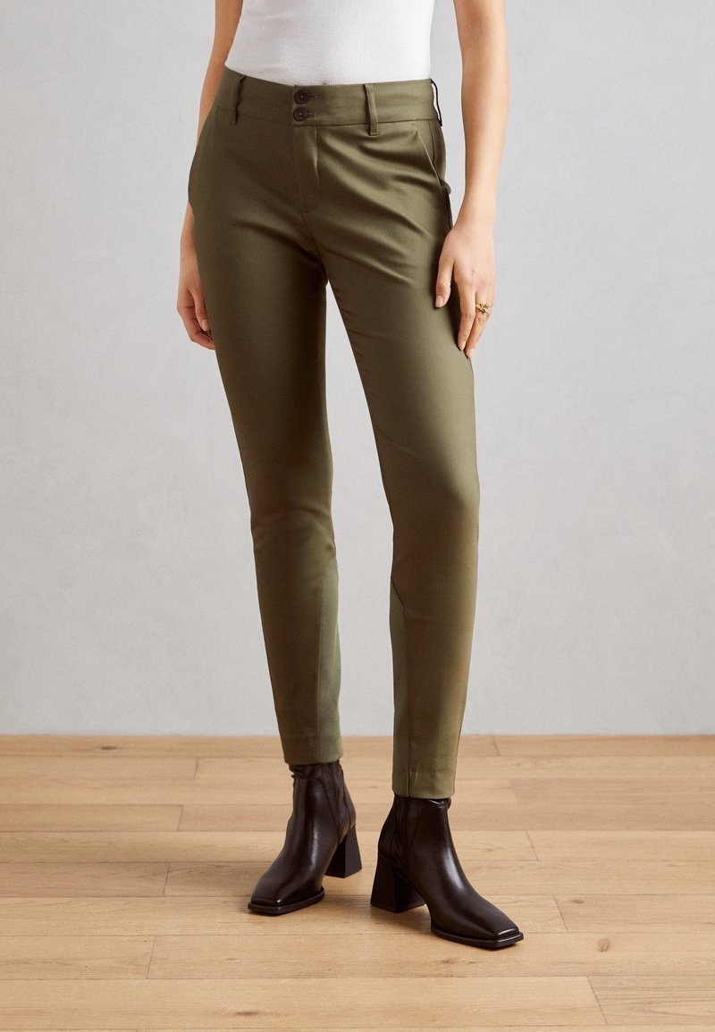 Pantalon slim vert olive à fermeture double bouton, porté avec des bottines noires à bout carré et un haut blanc sans manches.