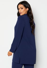 Tunic a maniche lunghe blu navy con una vestibilità rilassata, dotata di colletto, spacchi laterali e una texture liscia. La parte anteriore è più lunga di quella posteriore.