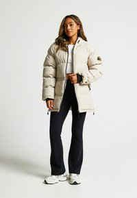 Bežová vypchávkovaná bunda s vysokým golierom, zipsom vpredu a elastickými manžetami. Skombinovaná s čiernymi rozšírenými nohavicami a bielymi teniskami.