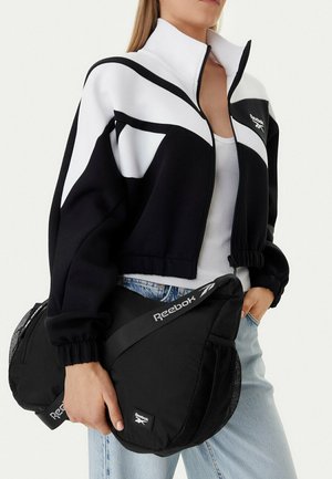 Mujer con chaqueta Reebok en blanco y negro sostiene dos bolsas negras de Reebok, vestida con top blanco y vaqueros azul claro.