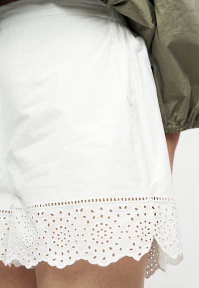 Λευκό σορτς με τελείωμα από δαντέλα με σκαλοπάτια που φοριέται από άτομο με πράσινο μακρυμάνικο πουκάμισο με το μανίκι διπλωμένο προς τα πάνω.