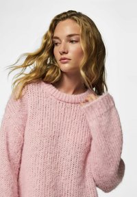 Jeune femme aux cheveux blonds ondulés portant un pull rose tricoté ample, regardant légèrement vers sa gauche sur un fond uni.
