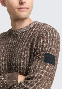 Brauner Kabelstrickpullover mit strukturiertem Muster, ausgestattet mit einem gerippten Rundhalsausschnitt und einem schwarzen Stoffpatch am linken Ärmel.