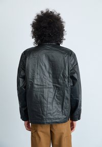 Giacca di pelle nera con una finitura liscia, vestibilità rilassata, polsini con bottone e lati con cerniera, indossata sopra pantaloni beige, vista da dietro.