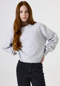 Grauer Pullover mit strukturierter Oberfläche, Rundhalsausschnitt und gerippten Bündchen. Kombiniert mit dunklen, gemusterten Jeans.