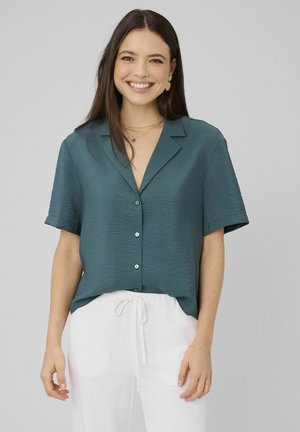 Femme souriante aux longs cheveux foncés porte une chemise turquoise à manches courtes boutonnée et un pantalon blanc à cordon, sur un fond gris uni.