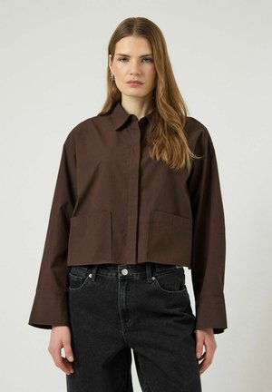 Femme aux longs cheveux châtain clair portant une veste boutonnée marron foncé et un jean noir taille haute, debout devant un fond clair uni.