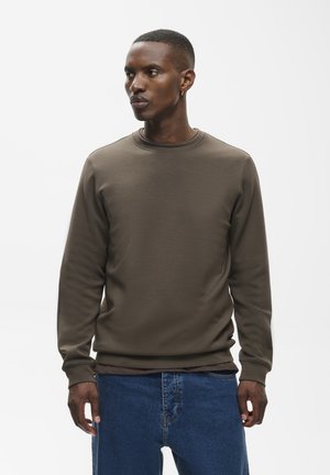 Jeune homme aux cheveux courts et portant une boucle d'oreille, habillé d'une chemise marron à manches longues et d'un jean bleu, regardant sur le côté sur un fond uni.