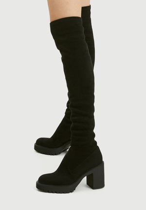 Black suede over-the-knee boots with chunky block heels and lug soles worn on bare legs against a plain background.
