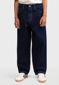 Jean large bleu foncé en denim, avec des coutures contrastées, une fermeture à bouton et des poches avant. Associé à des baskets blanches.