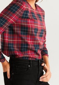 Persoon in een rood, marineblauw en wit geruit overhemd met lange mouwen, gestoken in hoge zwarte jeans, met de handen bij de zakken.