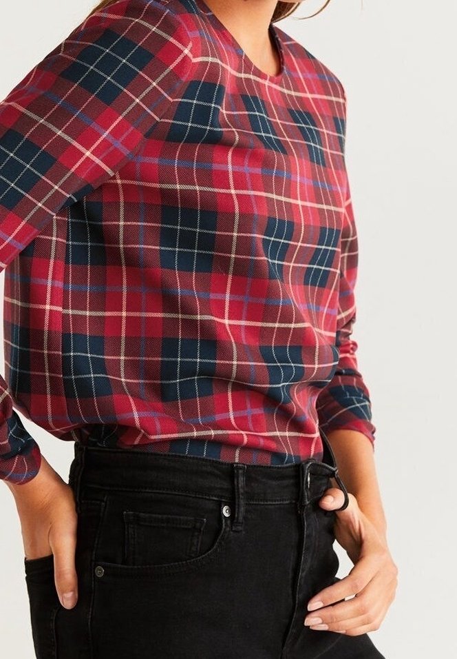 Persoon in een rood, marineblauw en wit geruit overhemd met lange mouwen, gestoken in hoge zwarte jeans, met de handen bij de zakken.