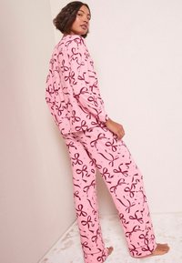 Rosa pyjamasset med lång ärm och vida ben, prydd med vinröda rosetter på mjukt tyg. Designen betonar komfort och lätthet.