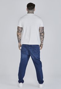 Valge lühikeste varrukatega T-särk, sinised teksapüksid lõdva istuvusega, kahe tagataskuga, ja valged tossud. Neutraalne taust.