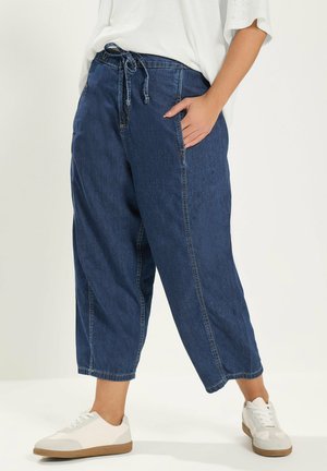 Person trägt lockere dunkelblaue, gekürzte Jeanshosen mit gebundener Taille und weißen Turnschuhen, steht mit einer Hand in der Tasche.