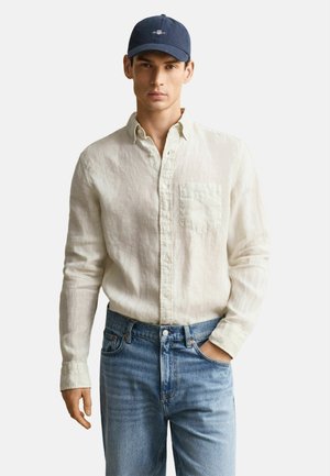 Junger Mann trägt ein beiges Hemd mit Knöpfen, blaue Jeans und eine marineblaue Baseballkappe, steht mit einer Hand in der Tasche.