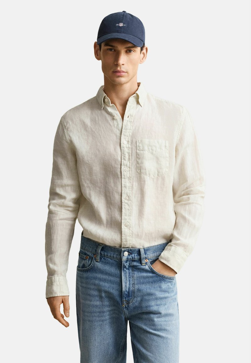 Jeune homme portant une chemise beige à boutons, un jean bleu et une casquette de baseball bleu marine, debout avec une main dans la poche.