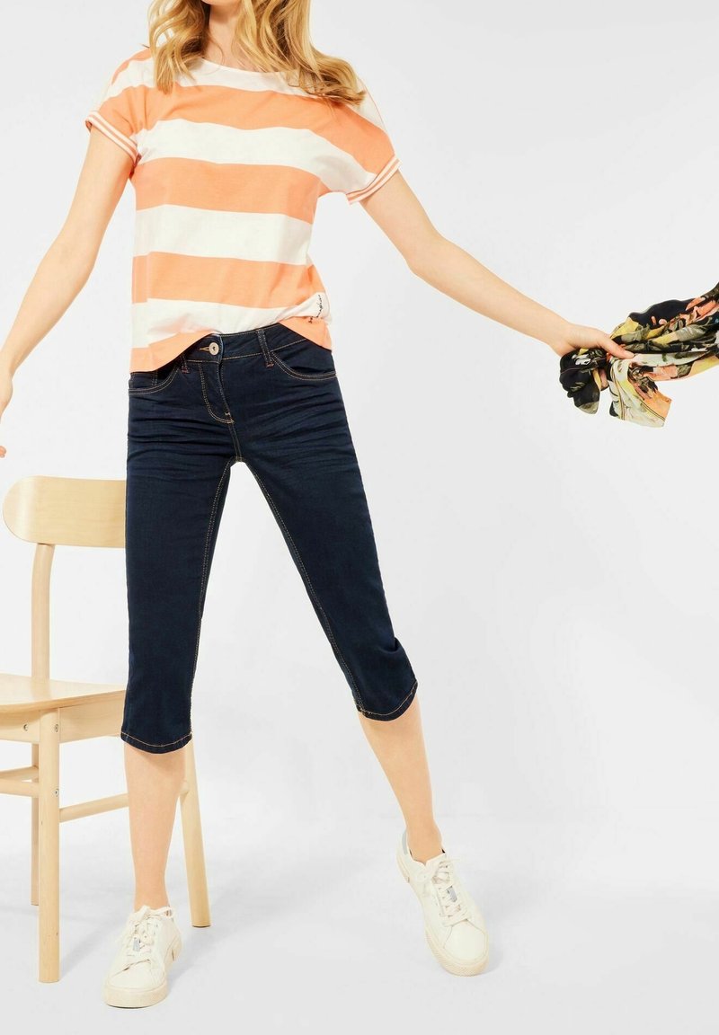 Oranssi-valkoraidallinen lyhythihainen paita, tummansiniset denim-caprishortsit ja valkoiset lenkkarit. Malli pitää kuvioitua huivia yhdellä kädellä.