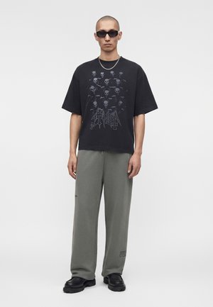 Άνδρας που φοράει μαύρη φαρδιά μπλούζα με σχέδιο κρανίου, γκρίζο παντελόνι με φαρδιά πόδια, μαύρα χοντρά παπούτσια, σκούρα γυαλιά ηλίου και ασημένιο κολιέ με αλυσίδα.