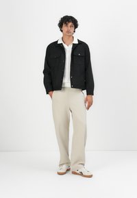 Zwarte suède jack met twee borstzakken, kraag en drukknopen, gedragen over een wit gebreid shirt en beige broek.