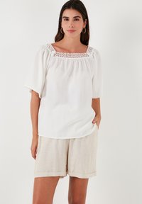 White blouse featuring a square neckline with lace trim, short sleeves, and a loose fit. Worn with beige linen shorts.