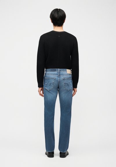 Άτομο που κοιτάζει προς τα πίσω, φοράει μαύρη ριμπ μακρυμάνικη μπλούζα, μπλε τζιν παντελόνι ίσιας γραμμής με ραφές στις πίσω τσέπες και μαύρα παπούτσια σε απλό φόντο.