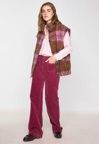 Gilet à carreaux multicolore en violet et jaune, à boutons. Assorti à un pantalon évasé en velours côtelé rose et des chaussures noires. Texture lisse.