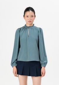 Teal bluse med volangkrage, lange puffermede ermer og en nøglehulsåpning foran. Kombinert med et marineblått plisserte skjørt.