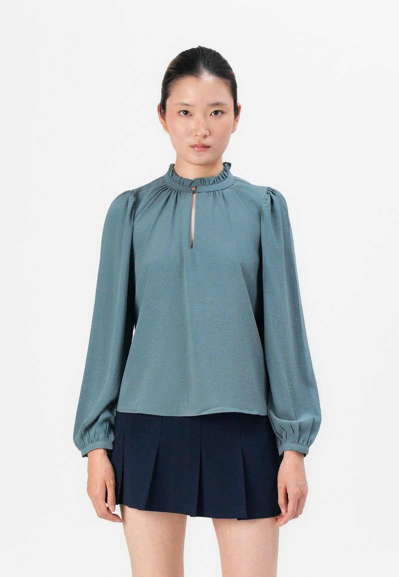 Teal bluse med volangkrage, lange puffermede ermer og en nøglehulsåpning foran. Kombinert med et marineblått plisserte skjørt.