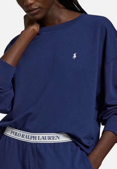 Γυναίκα με μπλε σκούρο μακρυμάνικο τοπ και παντελόνι που ταιριάζει, με λευκή ζώνη Polo Ralph Lauren, το χέρι της ακουμπάει κοντά στον λαιμό της.