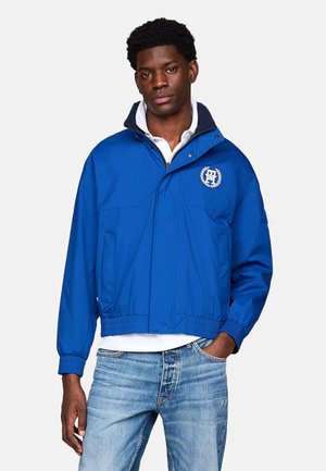Chaqueta azul, ligera, con capucha, puños elásticos, logo en el pecho y bolsillos frontales. Hecha de un material suave y resistente al agua.