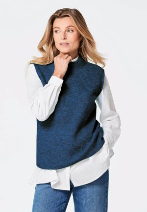 Top sans manches en maille bleu marine avec un col montant, superposé sur une chemise blanche, associé à un jean bleu. Le tissu présente une texture côtelée.