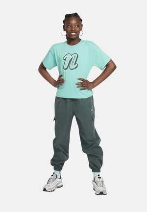 Chica sonriente de pie con las manos en las caderas, lleva una camiseta Nike azul claro, joggers verde oscuro, zapatillas plateadas y pendientes de aro.