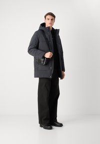 Puffy black parka with a hood, made of weather-resistant material, worn over a dark shirt, paired with wide black trousers and black boots.