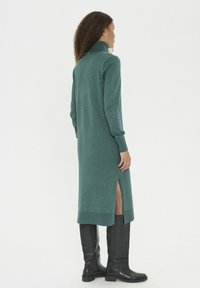 Femeie stând de profil, îmbrăcată cu o rochie tricotată lungă de culoare teal, cu guler înalt și o fante laterală, completată cu cizme negre până la genunchi.