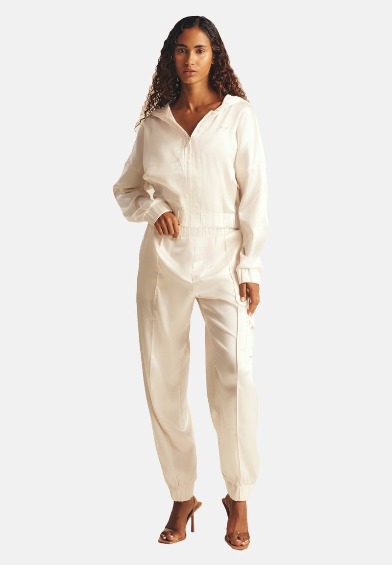 Ensemble de survêtement en satin blanc avec une veste à capuche zippée et un pantalon à taille élastique, comportant des poignets, des lignes fluides et une coupe décontractée.