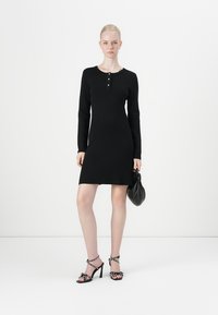 Black knit dress with long sleeves, round neckline, and three buttons. Model wears black heeled sandals with embellishments and a black bag.
