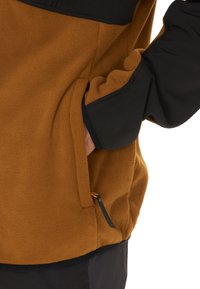Brun fleecejacka med en svart övre del, som har en ficka på sidan med en dekorativ dragkedja och en texturerad tygfinish.