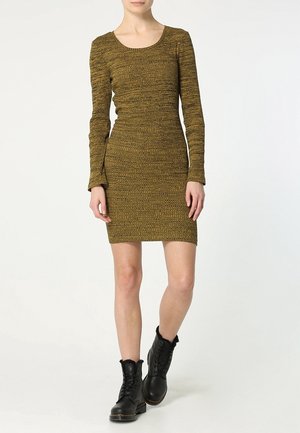 Femme portant une robe moulante texturée à manches longues de couleur vert olive et des bottines noires à lacets, debout sur un fond blanc.