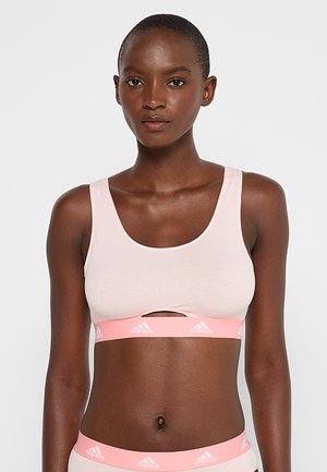 Woman wearing light pink sports bra and matching underwear with Adidas logo on elastic bands, standing against white background.
