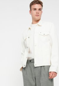 Fiatal férfi fehér gombos zakóban, fehér ingben és szürke kockás nadrágban áll, keze a zsebében, fehér háttér előtt.