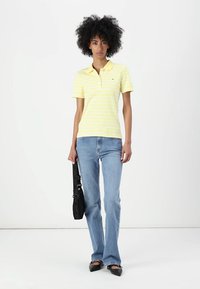 Gulstribet polo shirt med korte ærmer, parret med lyseblå vidde jeans. Modellen holder en sort taske med strop. Flade sorte sko.