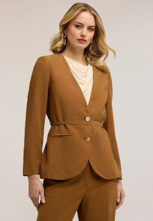 Vaaleahiuksinen nainen, jolla on suuret kultaiset rengaskorvakorut, ruskea istutettu bleiseri, jossa on napit, ja siihen sopivat housut neutraalia taustaa vasten.