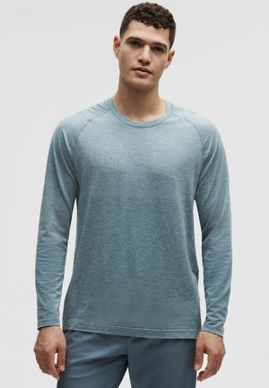 Ung mand iført en lyseblå langærmet tekstureret skjorte og matchende bukser, stående foran en ensfarvet lys baggrund.