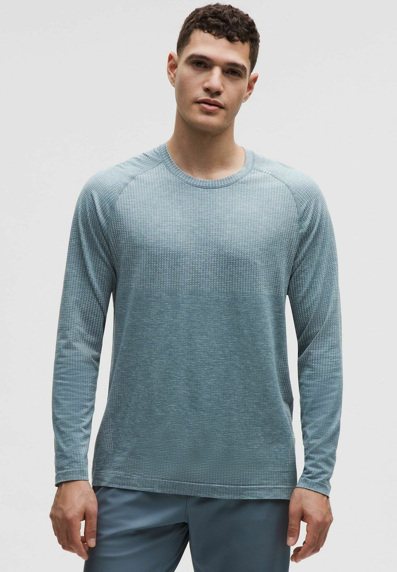 Mladi muškarac odjeven u svijetloplavu teksturiranu košulju dugih rukava i odgovarajuće hlače, stoji nasuprot jednobojnoj svijetloj pozadini.